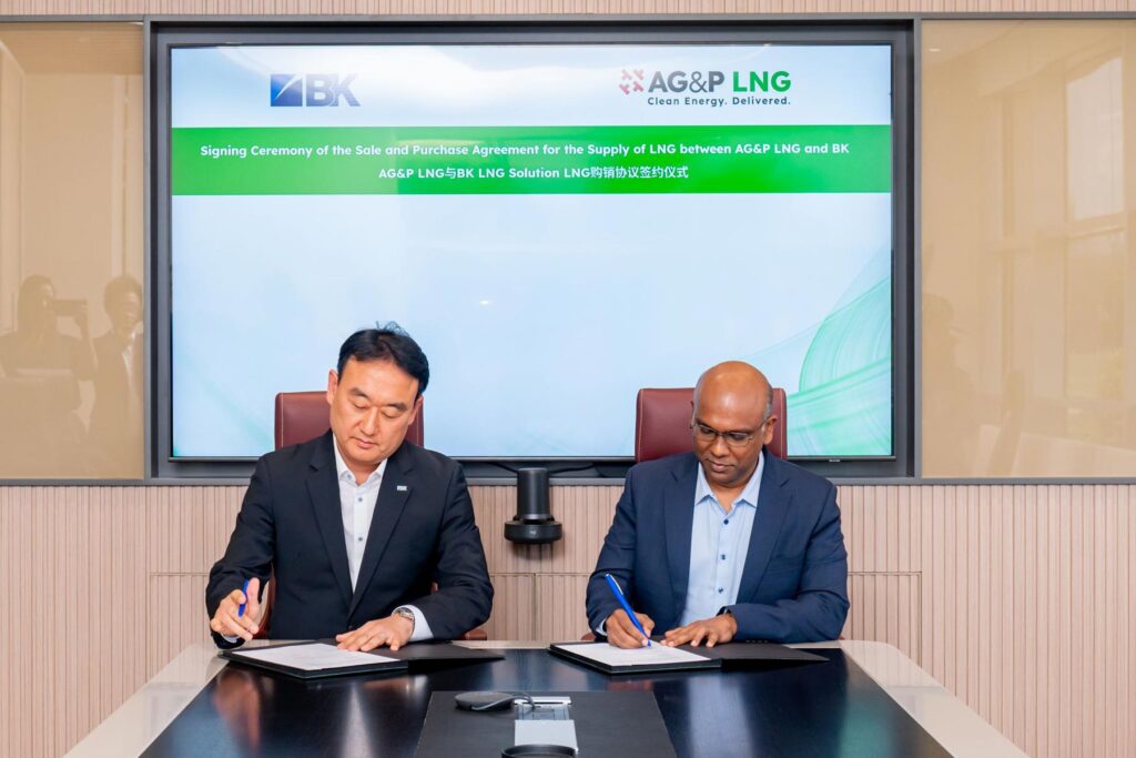 Two men sit at a table signing documents, with a screen displaying "Signing Ceremony of the Sale and Purchase Agreement for the Supply of LNG between AG&P LNG and BK" in the background.