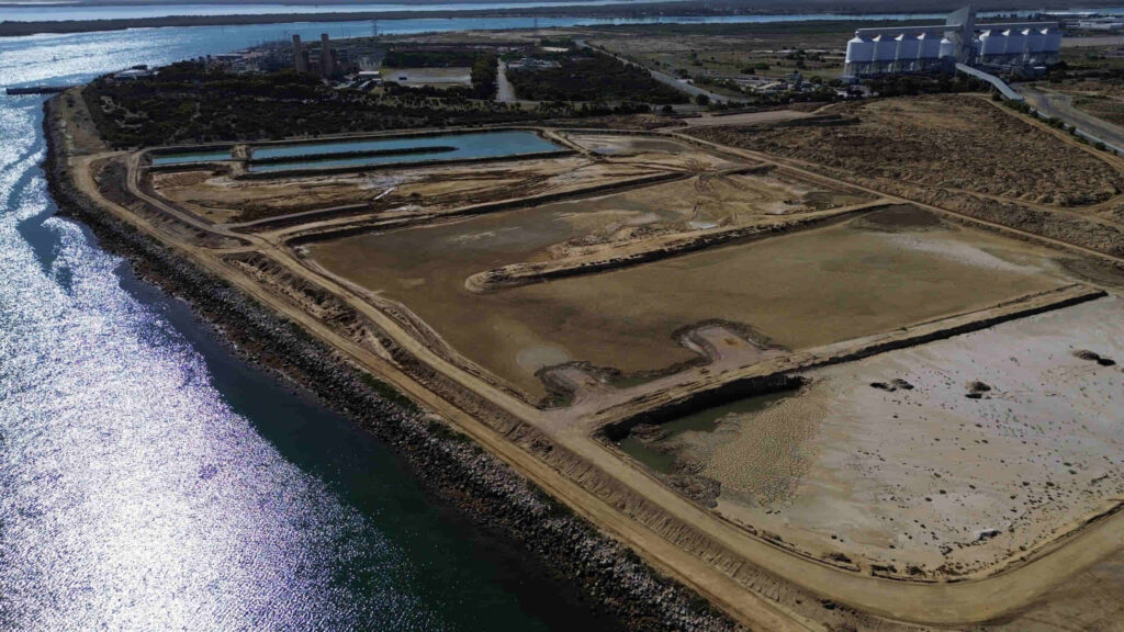 Aerial view of an industrial area with water channels, dirt roads, and storage buildings near a body of water.