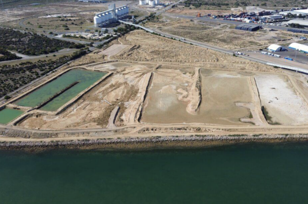 Aerial view of a coastal industrial area with large rectangular ponds, adjacent to a body of water and surrounded by sparse vegetation and roads.